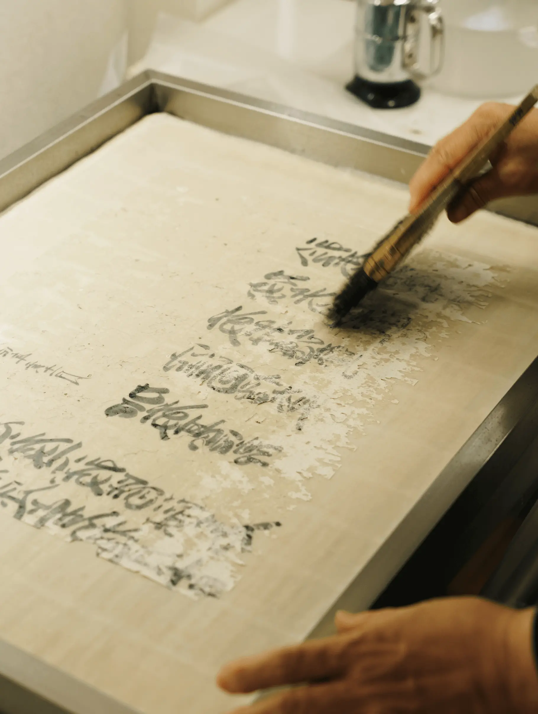 水槽の上で古文書を広げ、筆を使って紙の表面の汚れや劣化した繊維を丁寧に洗浄している修復作業の様子。株式会社辻村表具店（三重県伊勢市）による伝統的な古文書修復工程