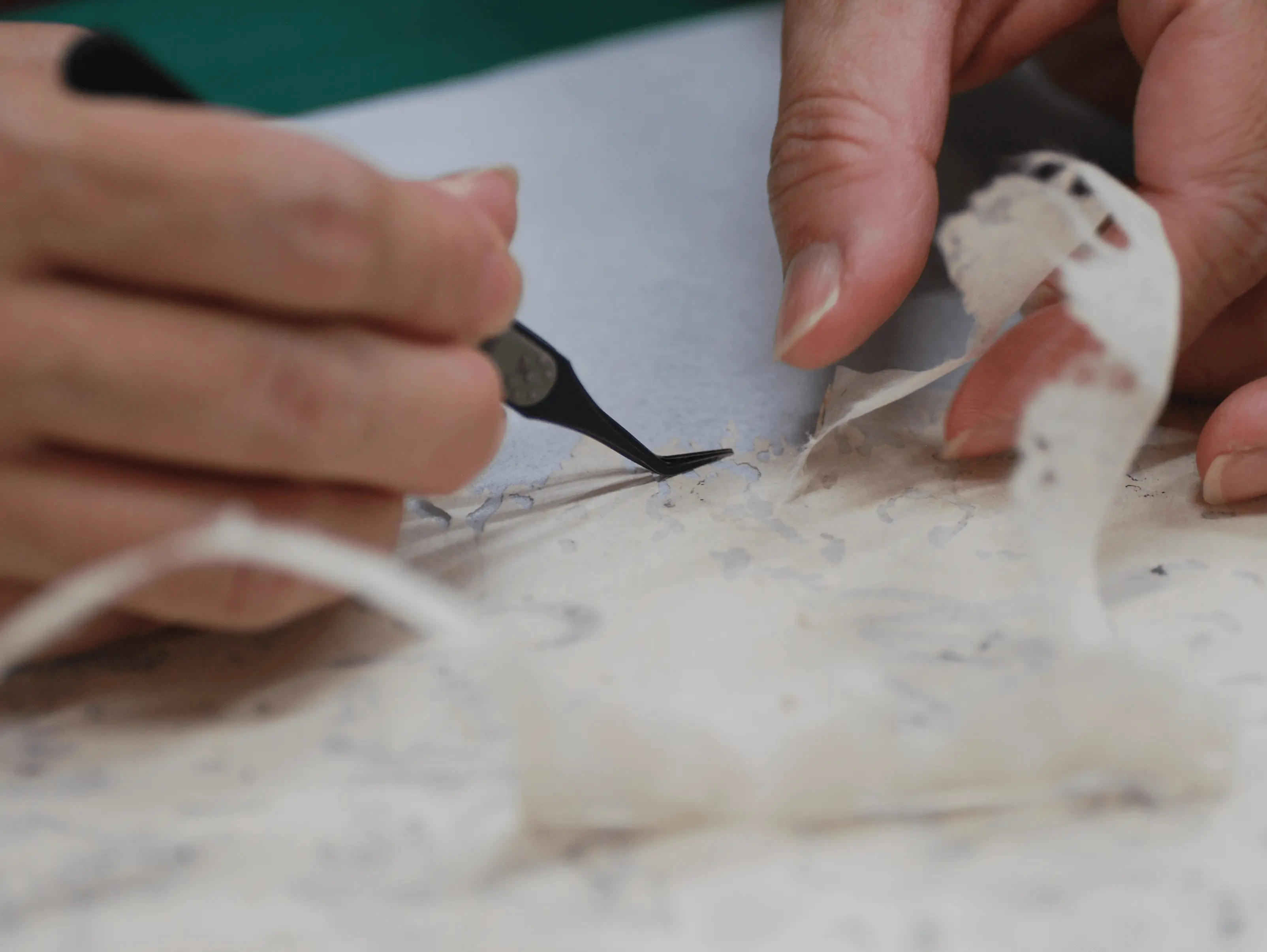 ピンセットを使って破れた古文書の繊維を整え、補修用の和紙を貼り重ねている細密な修復作業の手元の様子。三重県伊勢市の株式会社辻村表具店による古文書修復技術''