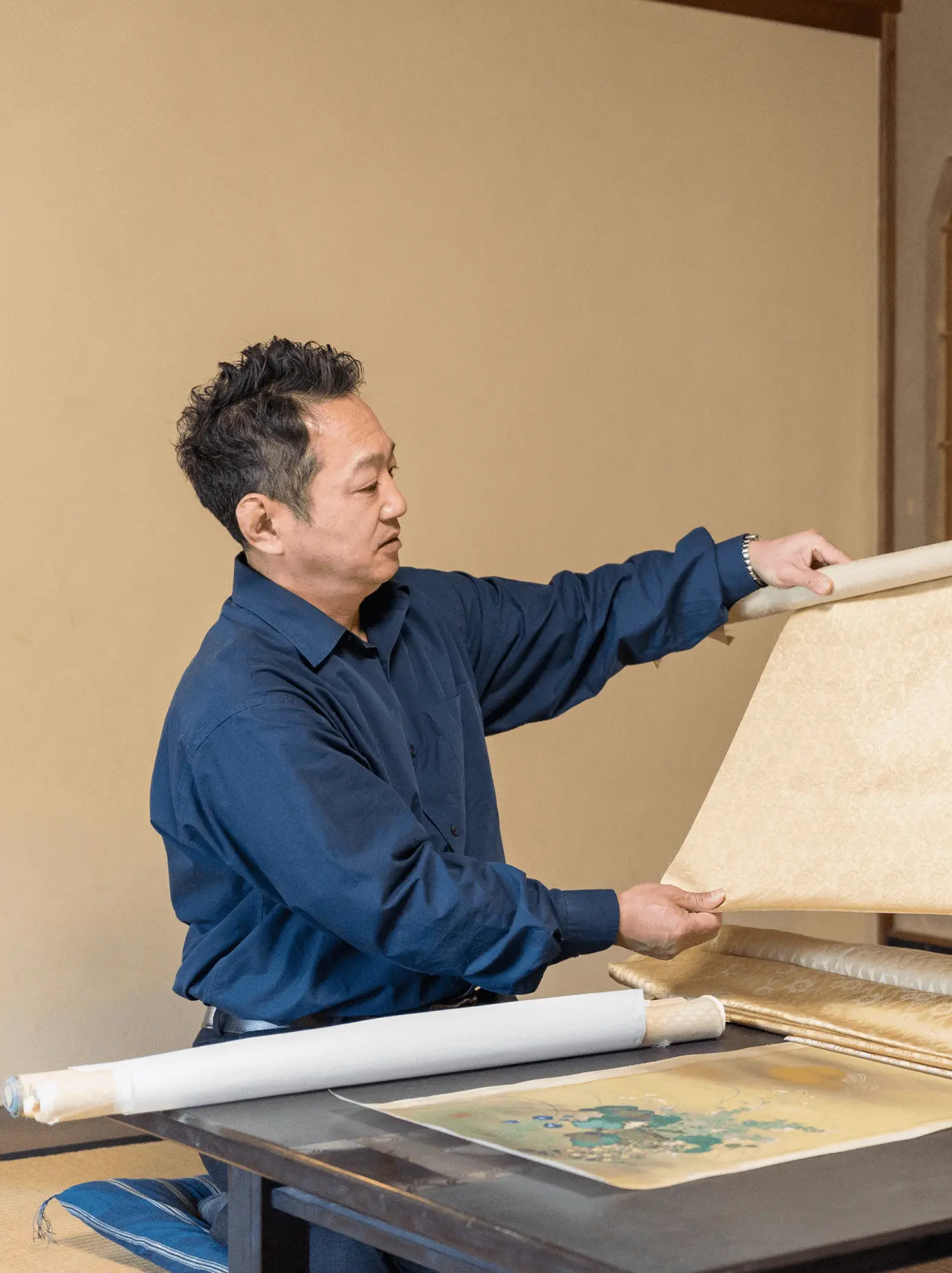 作業台の前で掛け軸の裂地を広げながら仕上がりを確認している表具師の様子。掛け軸修復や表装を行う三重県伊勢市の株式会社辻村表具店の職人による制作風景