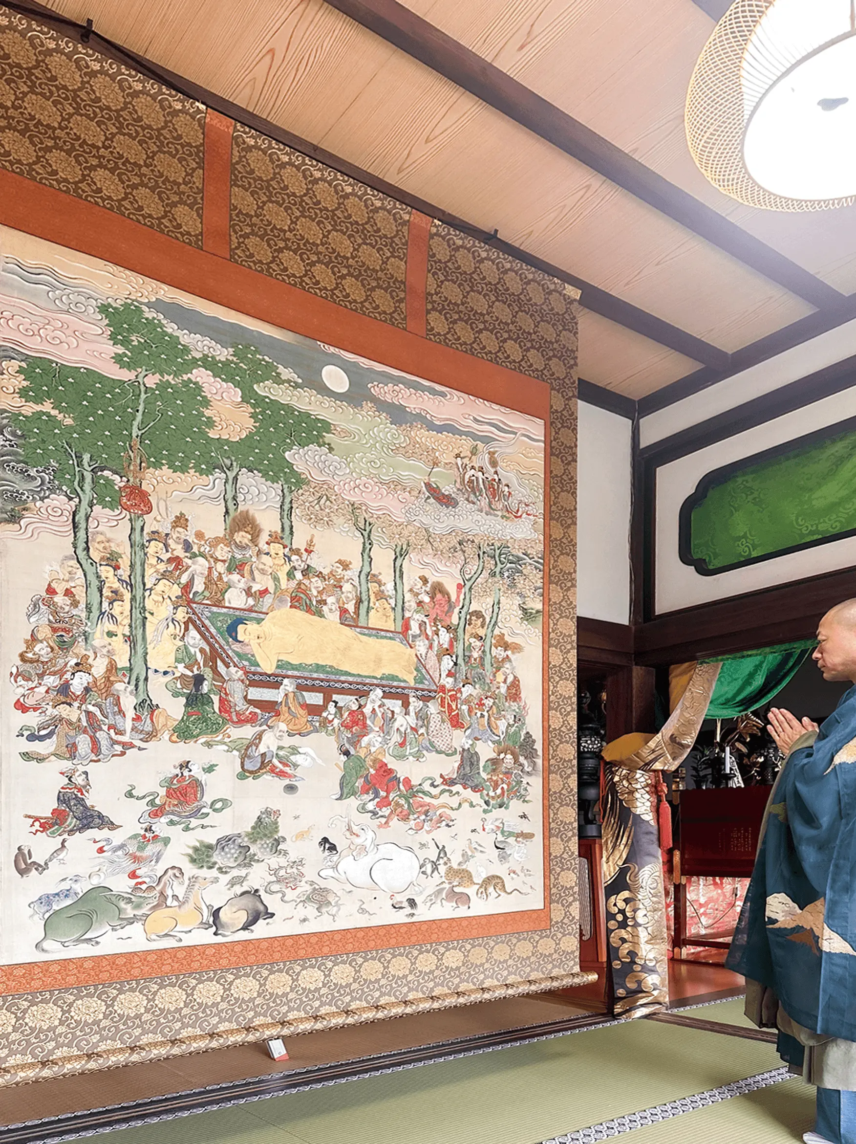寺院の本堂で大きな仏画の掛け軸が荘厳され、僧侶が合掌して拝んでいる様子。三重県伊勢市の株式会社辻村表具店が修復と表装を手がけた文化財掛け軸の納品後の風景