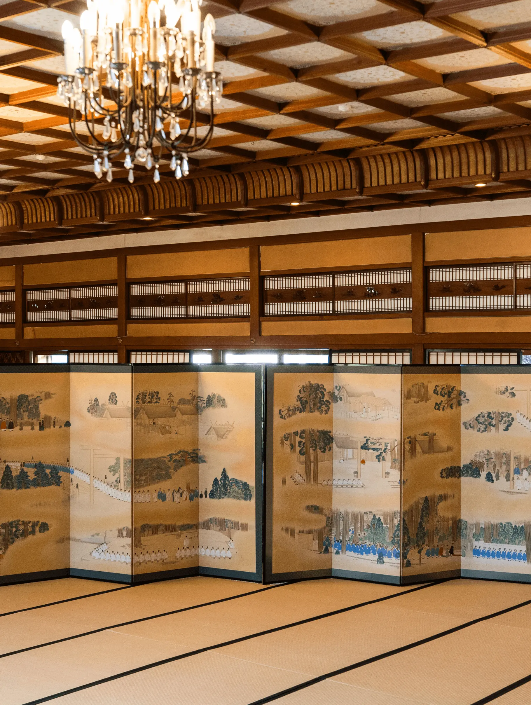 広い和室に設置された歴史風景を描いた6曲一双の屏風で、天井の格天井とシャンデリアがある空間で展示されている様子。三重県伊勢市の株式会社辻村表具店が修復と表装を手がけた屏風修復事例