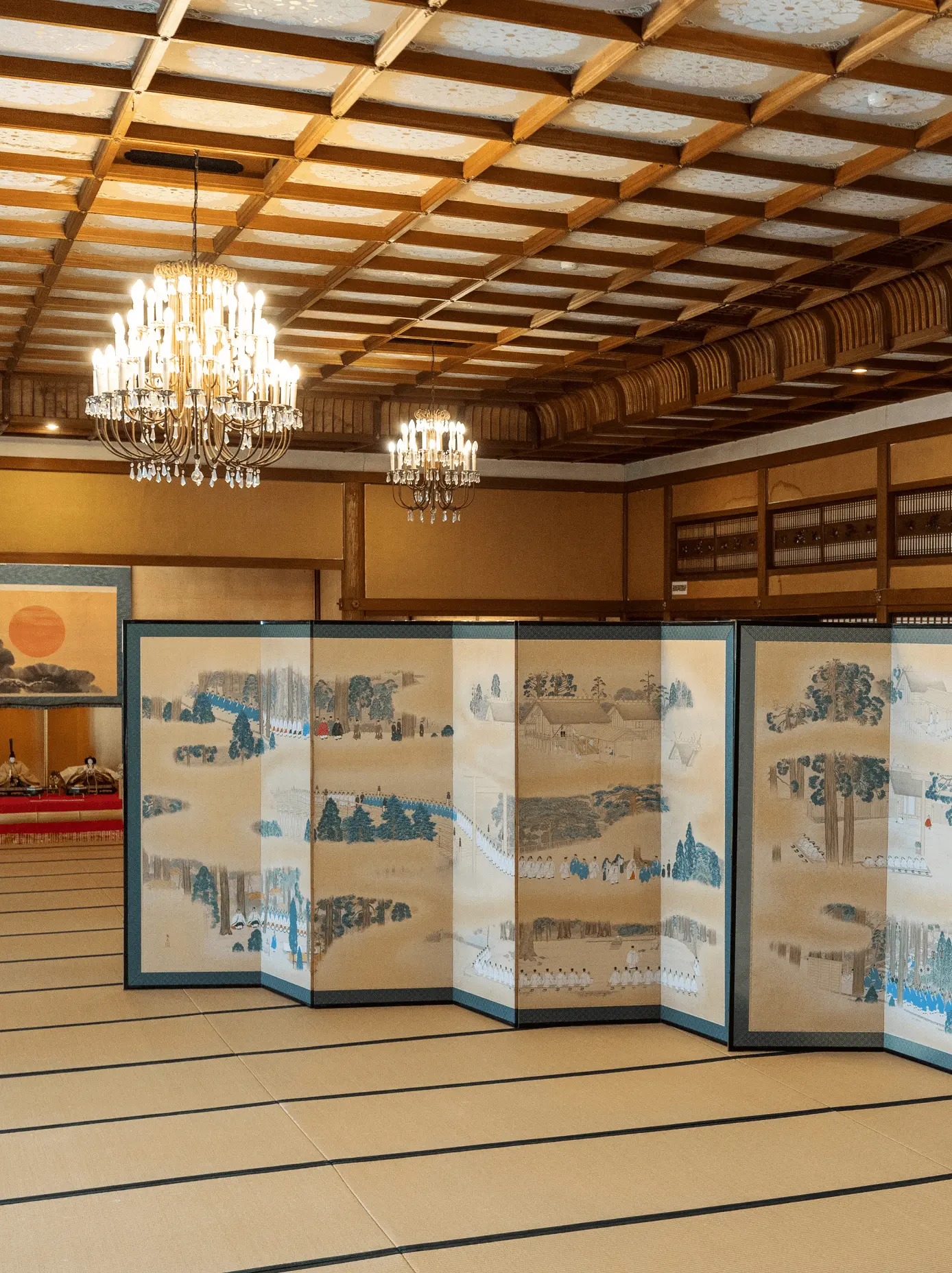 格天井とシャンデリアのある大広間に並べて設置された歴史絵巻風の屏風作品の全景。寺院や文化施設向けに屏風修復と表装を行う三重県伊勢市の株式会社辻村表具店の施工事例
