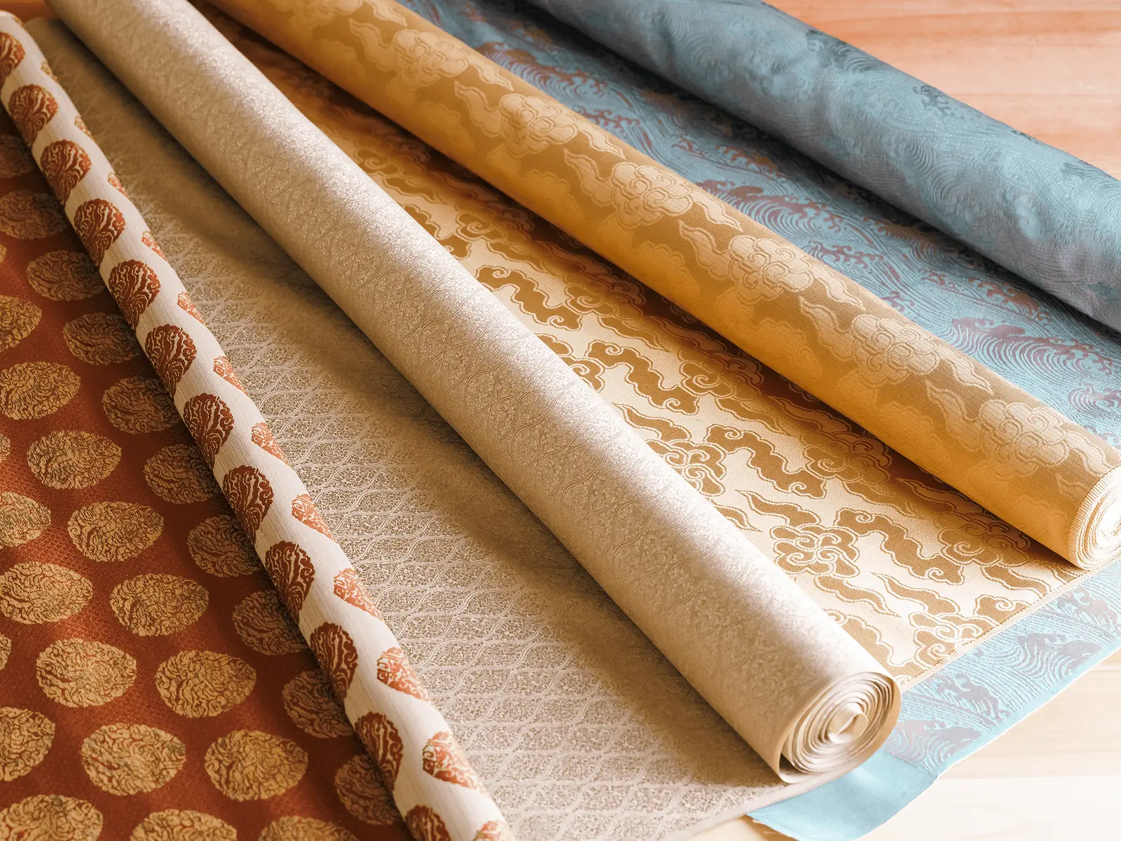 青や金、生成り、朱色など多様な文様の表装裂地が床に並べられており、掛け軸や襖の仕立てに用いる布地の種類と色合いを選定している工房の作業風景