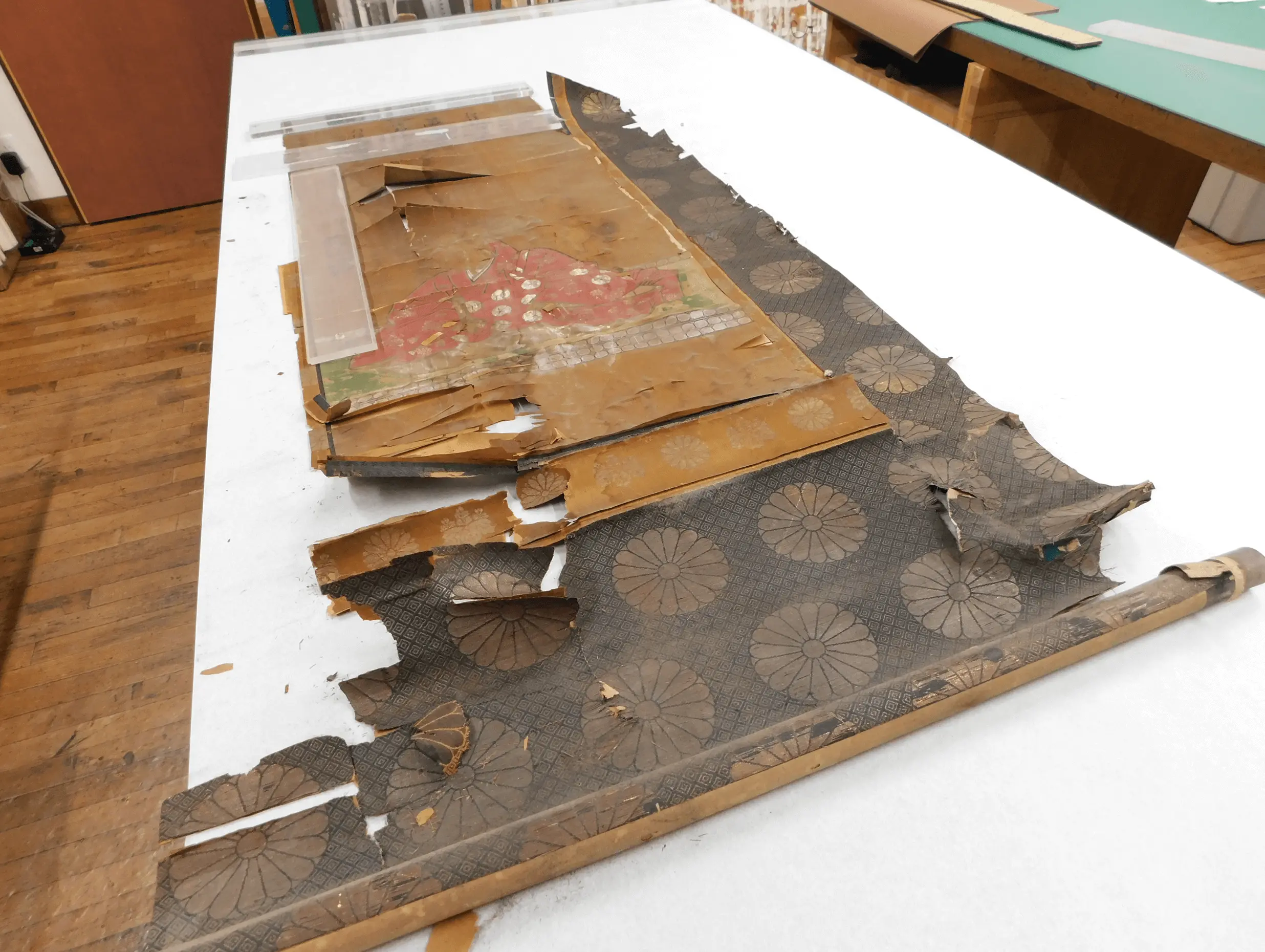 修復前の掛け軸を作業台の上に広げた状態。表装裂が大きく破れ、紙本も剥離や欠損が見られる古文書掛軸で、経年劣化による深刻な損傷が確認できる。掛け軸修復、文化財修理、古文書保存の施工前の様子。