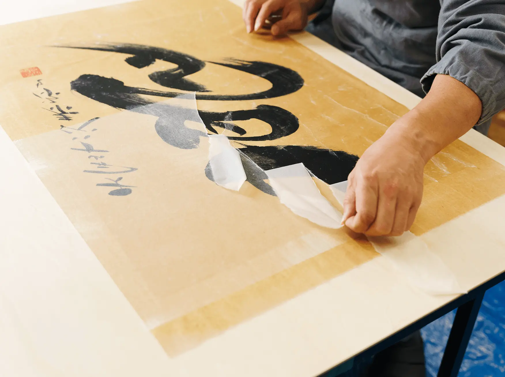 大きな書作品の表面に保護用の和紙を貼り重ね、墨書部分を傷めないよう丁寧に剥離作業を行っている表具修復工程の一場面。掛け軸や書画の修復・保存処理を行う職人の手元。