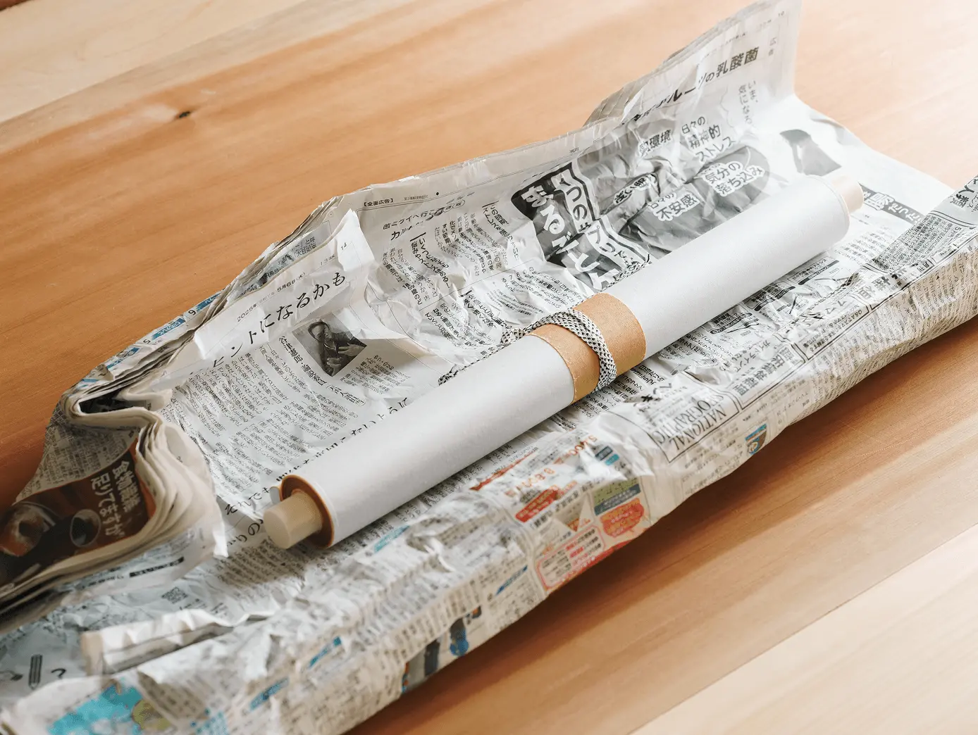 新聞紙で丁寧に包まれた掛け軸の保管状態。白い布で包んだ軸棒が新聞紙の中から見え、木製の台の上に置かれている適切な保管方法の実例