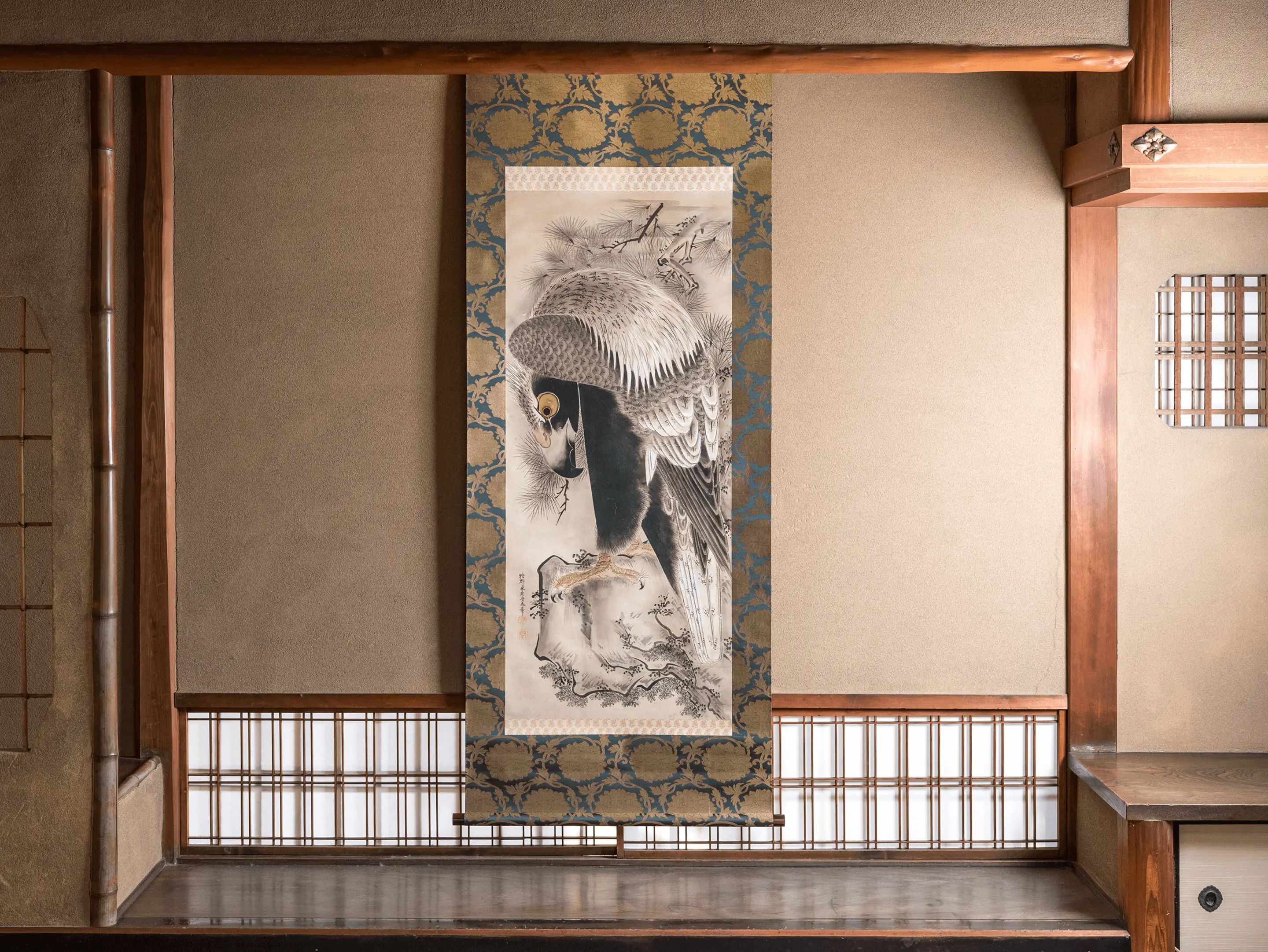 伊勢市の株式会社辻村表具店が保管する日本画の掛け軸。伝統的な和室の床の間に飾られた鷲と松を描いた墨絵の掛け軸。青と金色の唐草模様の表装が施され、格子戸のある和風建築の空間に調和している