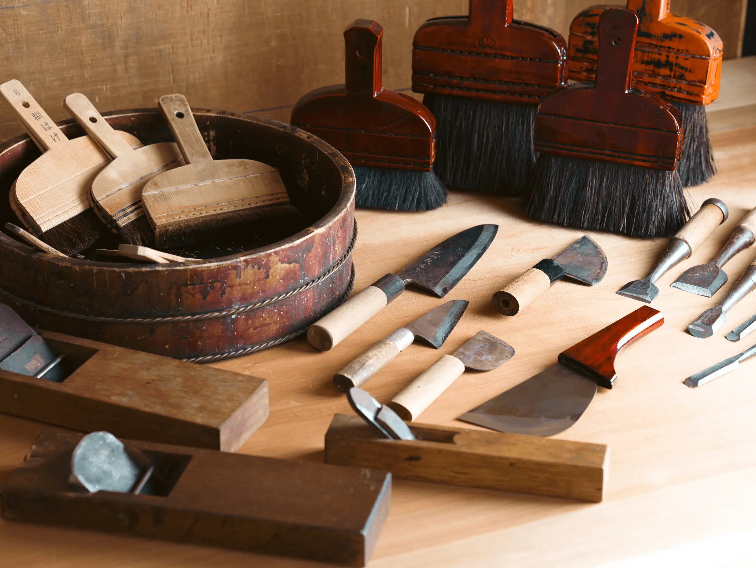 三重県伊勢市の株式会社辻村表具店の作業台に、裏打ち用刷毛や鉋、包丁など表具師の伝統工具が並べられており、掛け軸や古文書修復を行う専門工房の作業環境が伝わる
