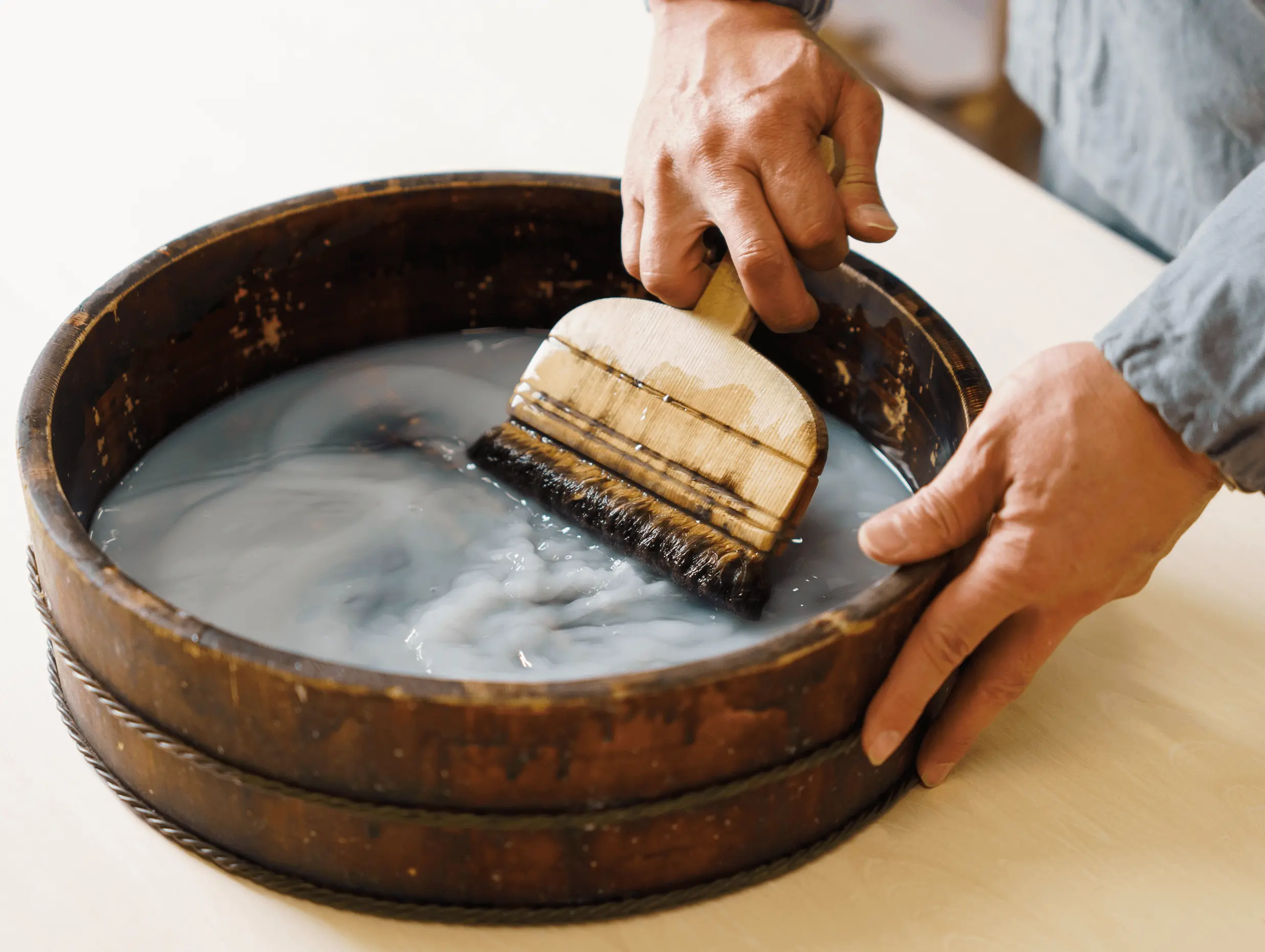 株式会社辻村表具店（三重県伊勢市）の職人が木桶に入った糊を裏打ち用刷毛ですくい取り、掛け軸の表装作業に使う準備をしている手元の様子