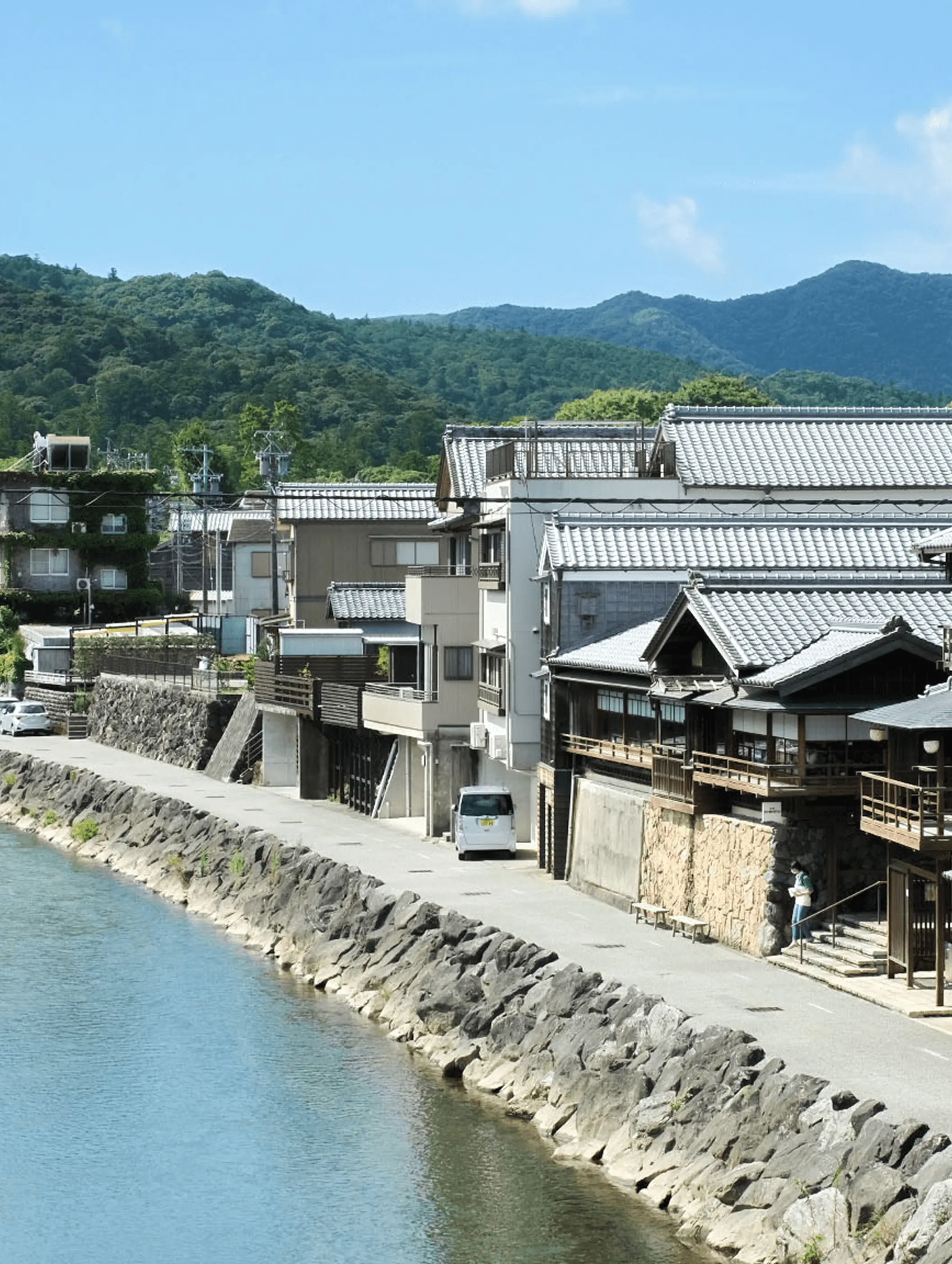 三重県伊勢市の五十鈴川沿いに並ぶ瓦屋根の町家や旅館の建物と山並みを望む風景で、伊勢の伝統的な町並みと落ち着いた観光地の雰囲気が分かる